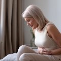 A woman in pain sitting on her bed clutching her stomach. 